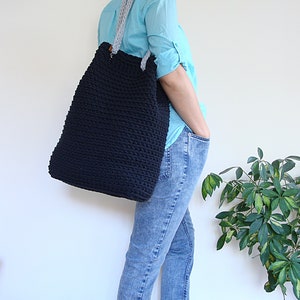 May include: A large black crocheted tote bag with gray straps. The bag is being worn by a person wearing blue jeans and a light blue shirt.