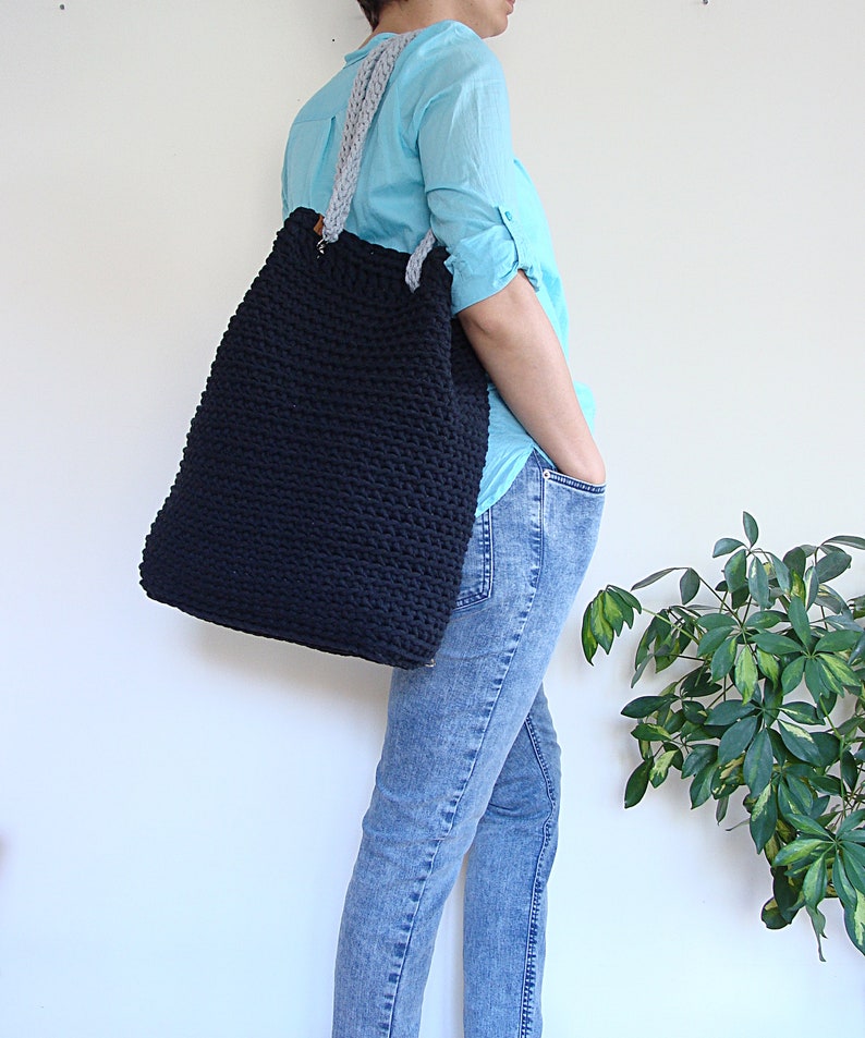 May include: A large black crocheted tote bag with gray straps. The bag is being worn by a person wearing blue jeans and a light blue shirt.