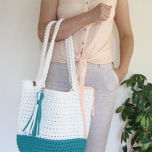 Puede incluir: Bolso tote blanco de crochet con fondo turquesa y un borla blanca. El bolso lo lleva una persona que lleva una camisa rosa claro y pantalones beige.