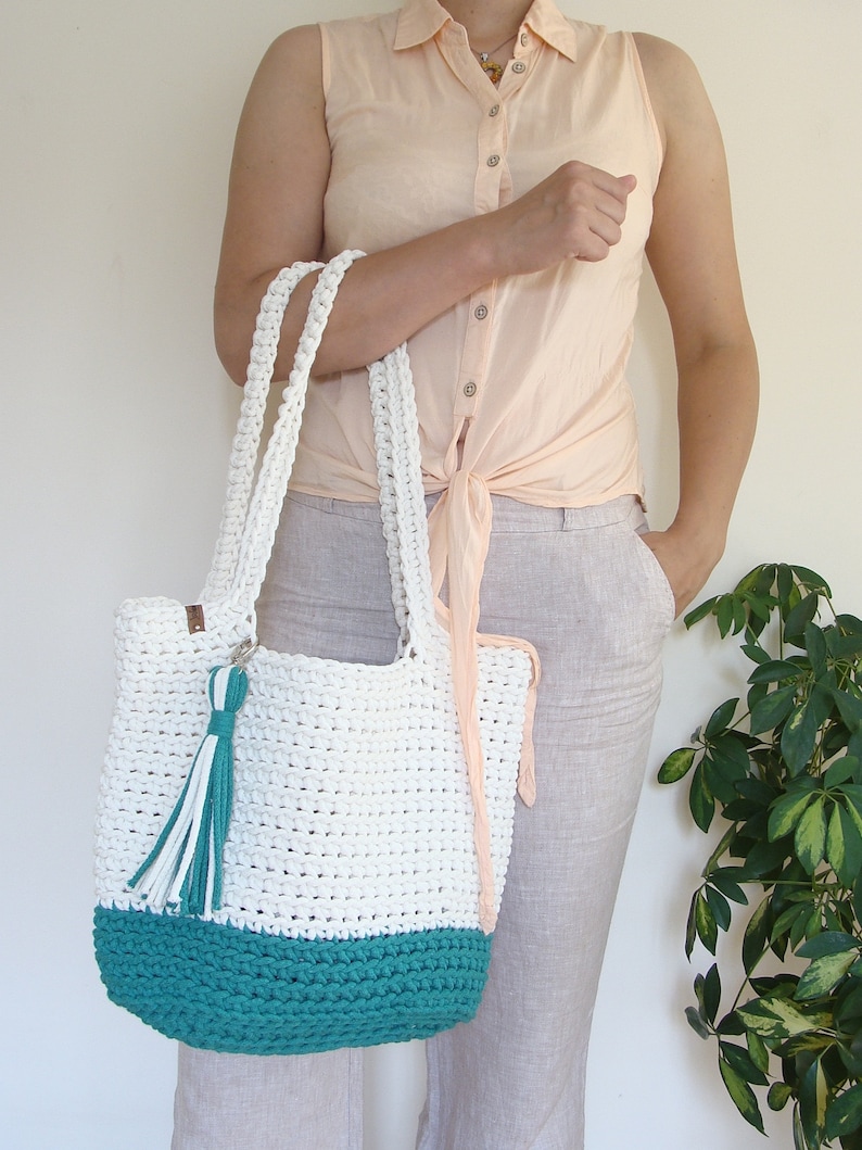 Puede incluir: Bolso tote blanco de crochet con fondo turquesa y un borla blanca. El bolso lo lleva una persona que lleva una camisa rosa claro y pantalones beige.