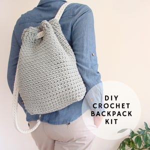 Può includere: Uno zaino all'uncinetto grigio chiaro con chiusura a coulisse e spallacci bianchi. Lo zaino è indossato sulla spalla. Il testo "DIY CROCHET BACKPACK KIT" è in un cerchio bianco.