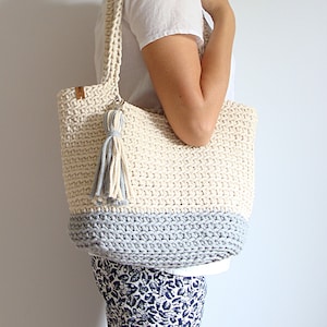Puede incluir: Un bolso tote de crochet con un borla gris y blanco. El bolso est&aacute; hecho de un hilo grueso y texturizado y tiene un fondo gris claro.