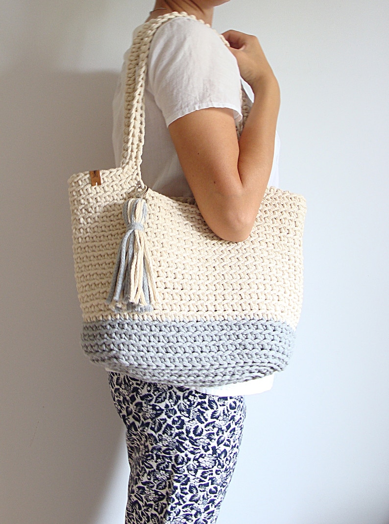 Puede incluir: Un bolso tote de crochet con un borla gris y blanco. El bolso est&aacute; hecho de un hilo grueso y texturizado y tiene un fondo gris claro.