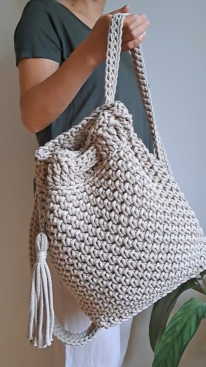 May include: A beige crocheted tote bag with a long strap and a tassel. The bag is made of thick, textured yarn and has a spacious interior.