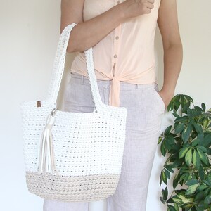Puede incluir: Un bolso tote de crochet blanco y beige con un borla. El bolso tiene una forma rectangular ancha y est&aacute; hecho con un hilo grueso y texturizado.