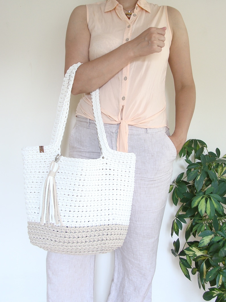 Puede incluir: Un bolso tote de crochet blanco y beige con un borla. El bolso tiene una forma rectangular ancha y est&aacute; hecho con un hilo grueso y texturizado.