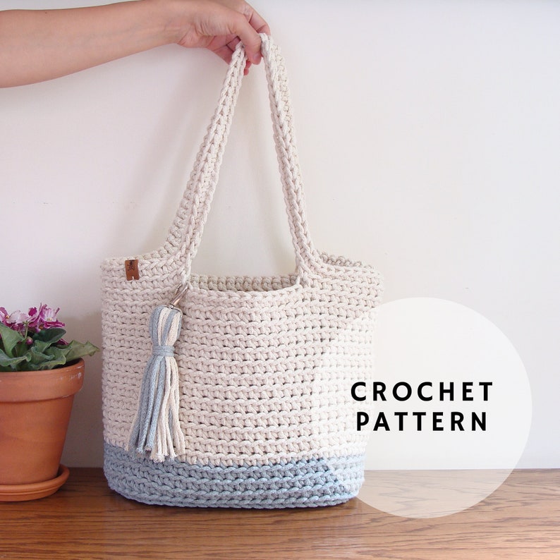 Puede incluir: Una bolsa de mano de crochet en color azul claro y blanco. La bolsa tiene un borl&oacute;n y el texto "CROCHET PATTERN" es visible en la imagen.