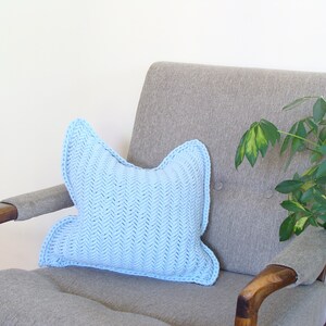 May include: A light blue knitted pillow with a chevron pattern sits on a gray armchair with wooden arms. A green plant is in the background.