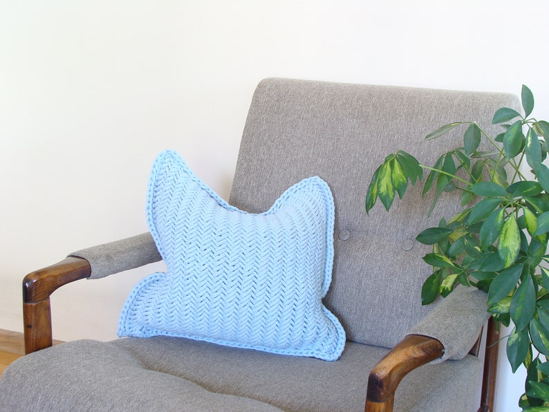 May include: A light blue knitted pillow with a chevron pattern sits on a gray armchair with wooden arms. A green plant is in the background.