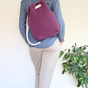 May include: A purple crocheted drawstring backpack with white straps. The backpack is being worn by a person.