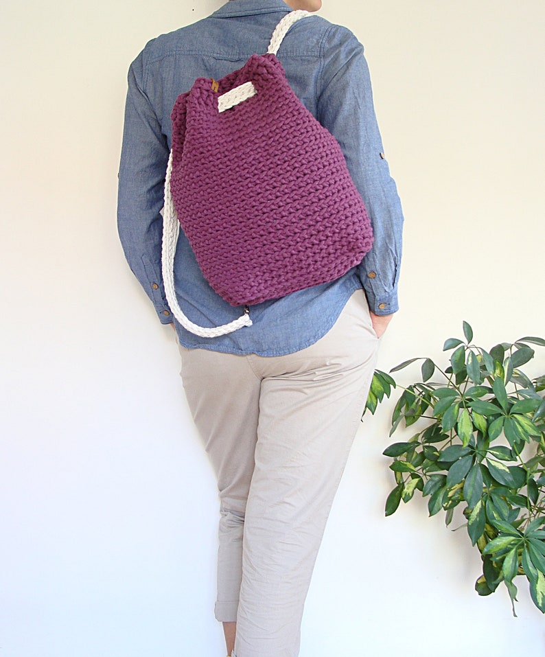 May include: A purple crocheted drawstring backpack with white straps. The backpack is being worn by a person.