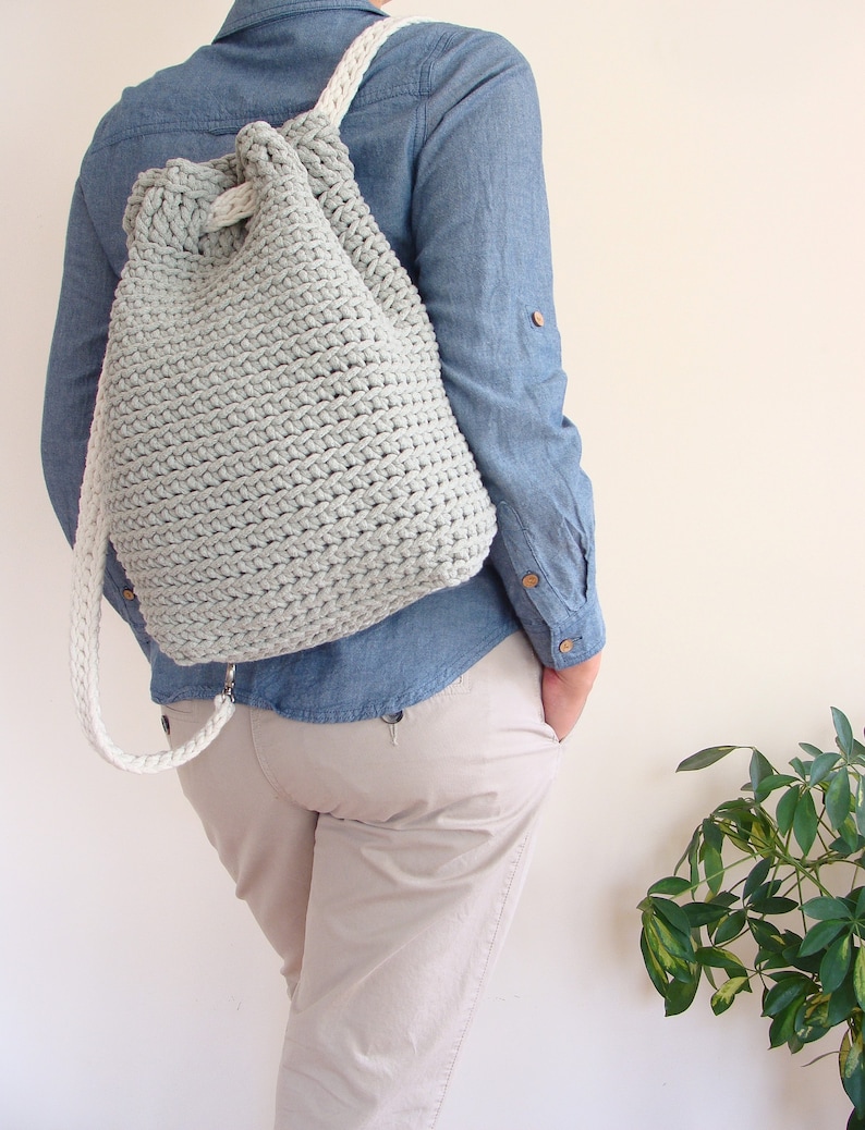 May include: A light gray crocheted drawstring backpack with white straps. The backpack is being worn by a person wearing a blue denim shirt and beige pants.