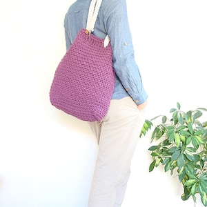 May include: A purple crocheted tote bag with white cotton straps. The bag is being worn by a person wearing a light blue shirt and beige pants.