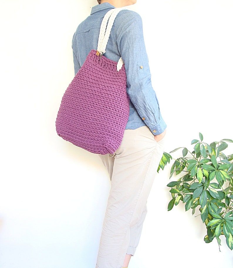 May include: A purple crocheted tote bag with white cotton straps. The bag is being worn by a person wearing a light blue shirt and beige pants.