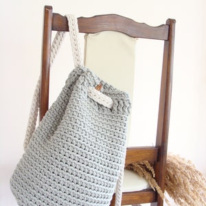 May include: A gray crocheted drawstring bag with white straps hanging on a wooden chair. The bag is made of thick yarn and has a simple, minimalist design.