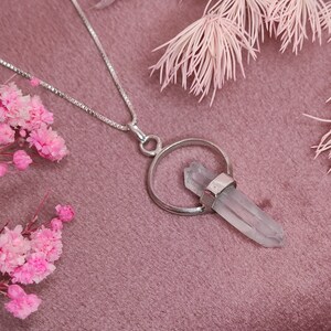 May include: A silver chain necklace with a clear quartz crystal pendant. The pendant is attached to a silver ring and the crystal is pointed at the top. The necklace is on a pink velvet surface.