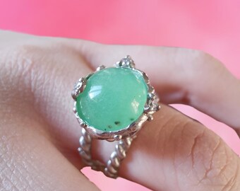 May include: A silver ring with a large, round, green gemstone set in a textured, silver setting. The ring is on a finger.