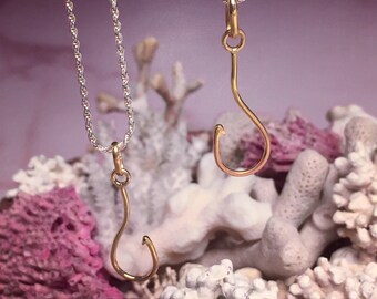 May include: Two gold fish hook pendants on silver chains. The hooks are hanging against a background of white and pink coral.