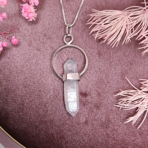 May include: A silver necklace with a clear crystal pendant. The pendant is shaped like a teardrop and is set in a silver circle. The necklace is on a pink velvet surface.