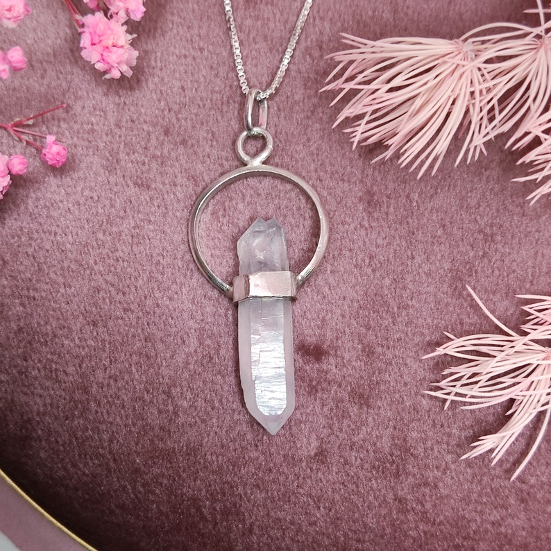 May include: A silver necklace with a clear crystal pendant. The pendant is shaped like a teardrop and is set in a silver circle. The necklace is on a pink velvet surface.