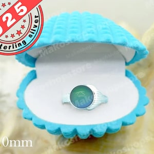 Pode incluir: Um anel de prata esterlina com uma pedra redonda que muda de cor é exibido dentro de uma caixa em forma de concha azul clara. A pedra do anel tem aproximadamente 10 mm de diâmetro. A caixa está aberta, revelando o anel.