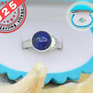 Pode incluir: Um anel de prata com uma pedra preciosa de lapis-lazúli azul engastada em um engaste de bisel. O anel é apresentado em uma caixa de anel de veludo azul.
