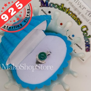 May include: A silver ring with a green gemstone set in a ring setting with a wing design. The ring is in a blue velvet ring box. The box is on a white surface with a colorful background that says "Moodstones Colors" and shows different colors of gemstones.