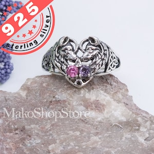 May include: Sterling silver ring featuring two wolf heads facing each other in a heart shape. The ring is adorned with three gemstones: two pink and one purple.