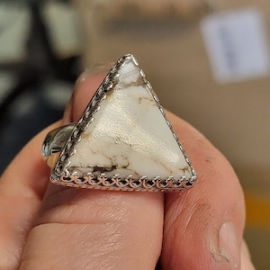 May include: A triangular ring with a white stone set in a silver bezel. The stone has a marbled pattern and is surrounded by a decorative silver border.