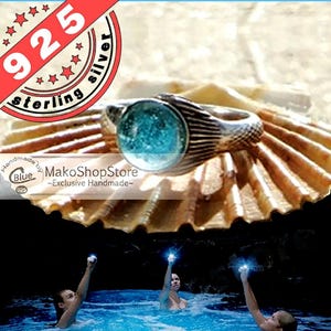 May include: Sterling silver ring with a blue gemstone and a mermaid tail design. The ring is displayed on a seashell. The text "925 sterling silver" is in the upper left corner of the image.