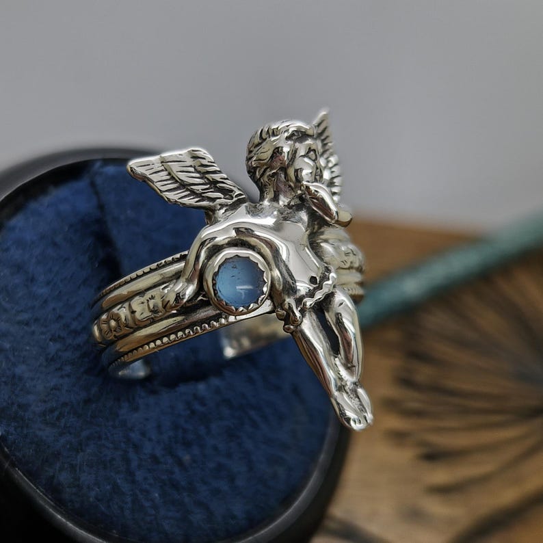 May include: A silver ring featuring a detailed cherub with outstretched wings. The ring has a light blue gemstone and intricate detailing around the band. The cherub is sculpted in a sitting position, adding a unique touch to this jewelry piece.