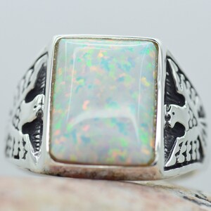 Puede incluir: Un anillo de plata con una piedra de ópalo blanco engastada en un bisel rectangular. El anillo presenta un diseño intrincado de dos pájaros a cada lado de la piedra.