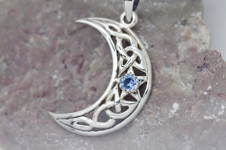 K&ouml;nnte beinhalten: Ein silberner Halbmond-Anh&auml;nger mit einem blauen Edelstein in der Mitte. Der Mond ist mit einem keltischen Knotendesign verziert.