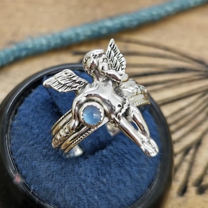 May include: A silver ring featuring a detailed cherub design with outstretched wings and a small, round, light blue gemstone. The ring is set against a dark blue velvet background.