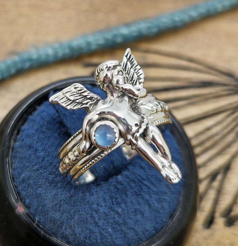 May include: A silver ring featuring a detailed cherub design with outstretched wings and a small, round, light blue gemstone. The ring is set against a dark blue velvet background.