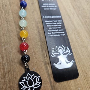Könnte beinhalten: Ein schwarzes Lesezeichen mit dem Wort "Chakra" und einer Liste der sieben Chakren mit ihren entsprechenden Farben und Positionen. Das Lesezeichen ist an einer Perlenkette mit sieben Perlen befestigt, die die Chakren repräsentieren. Die Perlen sind lila, blau, grün, hellgrün, gelb, orange und rot. Das Lesezeichen zeigt auch ein weißes Lotusblüten-Design.