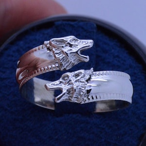 May include: A silver ring with two wolf heads facing opposite directions. The ring is made of polished silver and has a textured band.