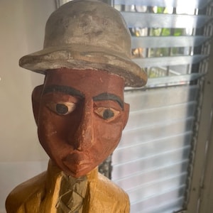 Op de afbeelding: Een houten beeldje van een persoon die een grijze hoed en een geel jack draagt. Het beeldje heeft een bruin gezicht met geschilderde kenmerken.