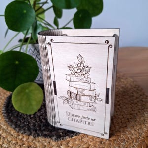 Peut inclure: Boîte en bois en forme de livre, avec un motif gravé au laser de livres empilés et de fleurs. La boîte est de couleur claire, avec une tranche incurvée et le texte français "Encore juste un CHAPITRE".
