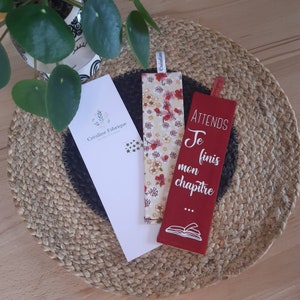 Peut inclure: Deux marque-pages en tissu rouge et blanc. Un marque-page a un motif floral et l'autre a le texte "ATTENDS Je finis mon chapitre..." avec une illustration de livre.