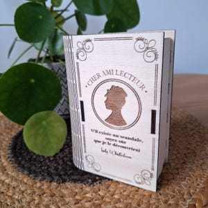 Peut inclure: Boîte en bois en forme de livre, avec texte gravé et silhouette de femme. Le texte dit "Cher ami lecteur" et une citation de Lady Whistledown. La boîte est de couleur claire avec des motifs décoratifs.