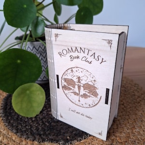Peut inclure: Une boîte en bois clair en forme de livre avec un motif gravé au laser. La couverture présente le texte "ROMANTASY Book Club" et "I will not die today" entourant une illustration circulaire d'un dragon volant au-dessus des nuages et des étoiles.