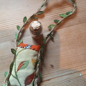 Puede incluir: Una pequeña bolsa de tela beige con un diseño floral, acentuada con un ribete naranja, está suspendida por una guirnalda de cuerda y hojas. Una pequeña botella de vidrio con un tapón en forma de flor está cerca. Los artículos están dispuestos sobre una superficie de madera.