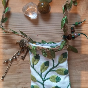 May include: A small white pouch with green leaf embroidery, a rope and leaf trim, and a key charm. The pouch is adorned with a rope handle and decorative leaves. Three stones are placed near the pouch.