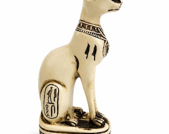 May include: An off-white cat figurine in an Egyptian style, featuring dark brown etched details including a decorative collar, hieroglyphic symbols on the side, and a patterned base. The cat is depicted in a seated, upright posture.