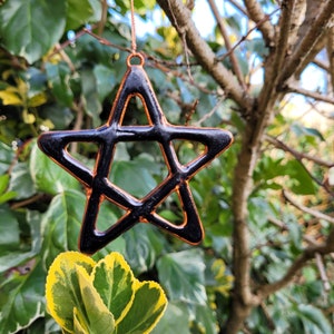 May include: A black pentagram star ornament with a copper finish hanging from a branch. The star is a five-pointed star with a copper outline and a black interior.