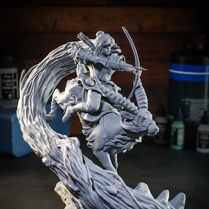May include: A gray 3D printed figurine of a woman riding a deer-like creature. The woman is holding a bow and arrow and is in a dynamic pose. The base of the figurine is a swirling, tree-like structure. The figurine is made by Nom Nom Figures.