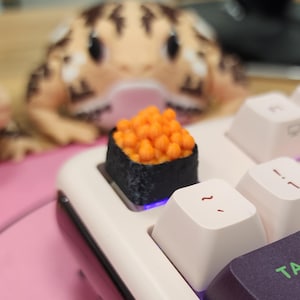 May include: A close-up of a sushi-shaped keycap with orange roe on a white keyboard. The keycap is black with orange details. Two gecko plush toys are in the background, out of focus.