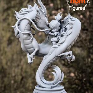 K&ouml;nnte beinhalten: Eine graue, detaillierte Figur eines Drachen und einer kleinen menschlichen Figur. Der Drache ist um den Menschen herumgewickelt, und die beiden Figuren ber&uuml;hren sich mit der Nase. Die Figur steht auf einem runden Sockel mit einem dekorativen Design.