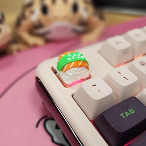 May include: A close-up of a keyboard with a sushi-shaped keycap. The keycap is white, orange, and green, resembling a piece of sushi. The keyboard has white keys and a black tab key with green text. A pink surface is in the background.